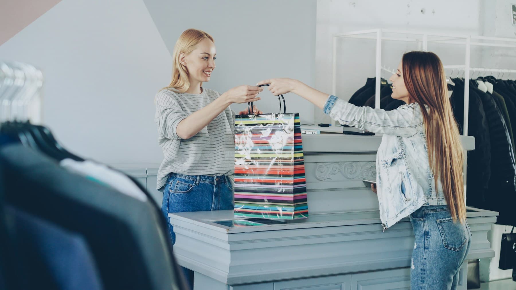 Two smiling women exchanging a shopping bag at a clothing-store counter